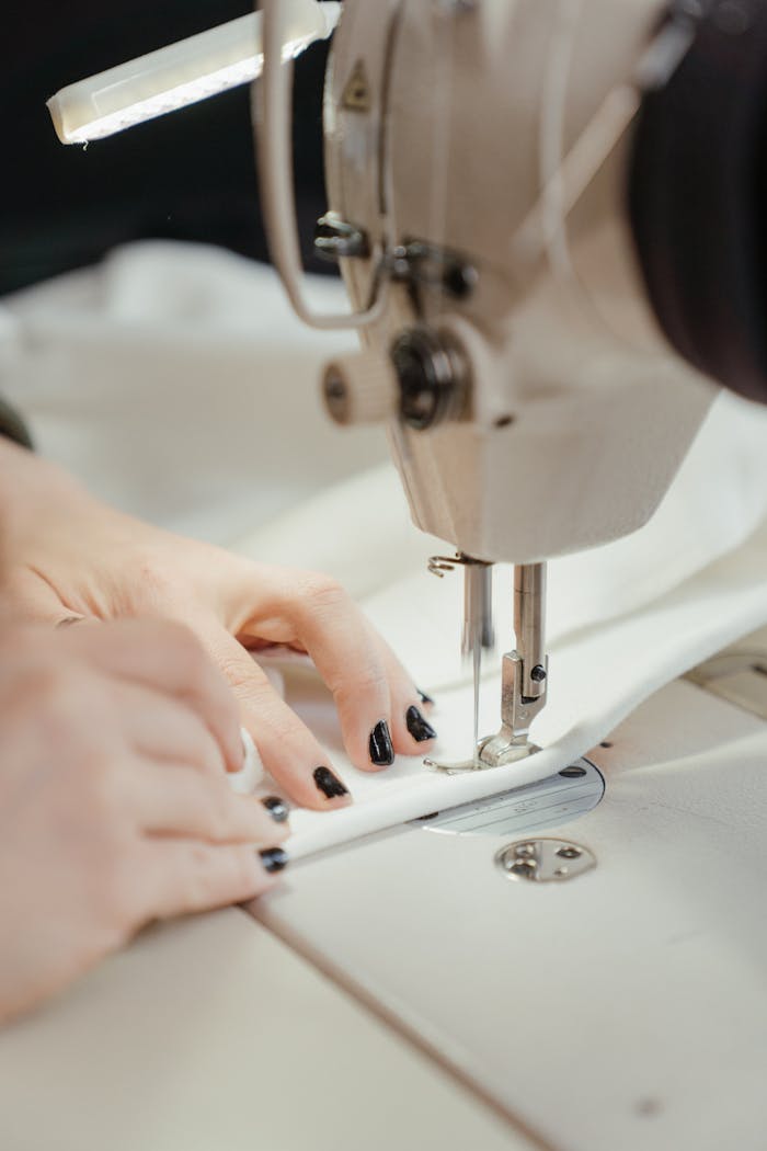 services-img Detailed view of a sewing machine stitching fabric with visible hands, nails painted black.